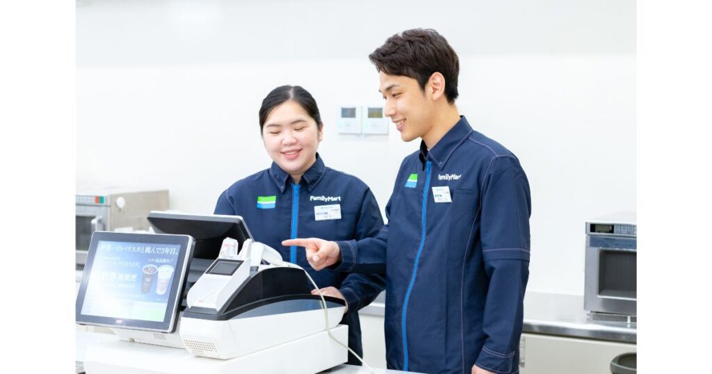 ファミリーマートのユニフォームを着た2人の従業員が、セルフレジの前で男性従業員が女性従業員に操作方法を笑顔で説明している写真。