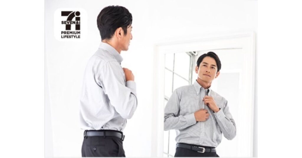 「Seven & i PREMIUM LIFESTYLE」のパッケージを手に、鏡の前でグレーのシャツを試着する男性。