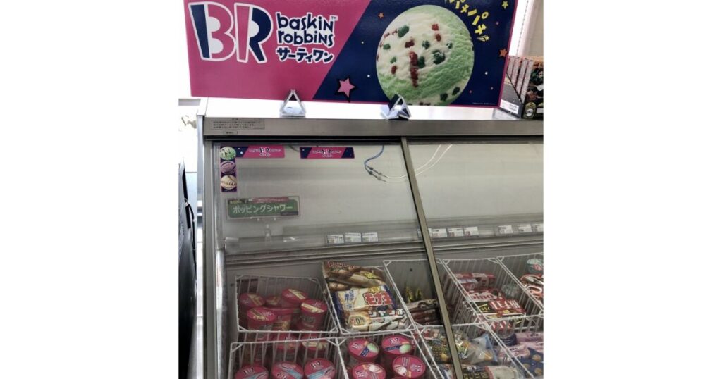 日本のコンビニ店内に置かれた、サーティワンアイスクリーム専用の冷凍ショーケース。上部のピンク色の看板にはBRのロゴと、ポッピングシャワーアイスの画像がある。冷凍庫の中には、BRのカップアイスが並ぶバスケットが見える。