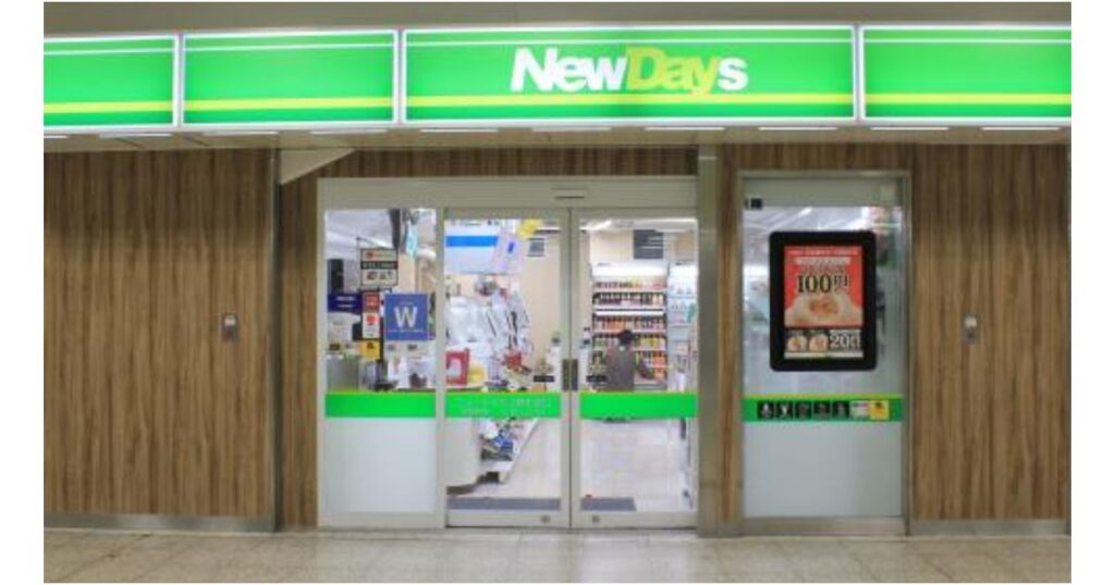 NewDaysコンビニエンスストアの店舗外観。緑色の看板に「NewDays」の白い文字。ガラスドアと木目調の壁。右側のデジタルサイネージには日本語のポスターが表示されている。