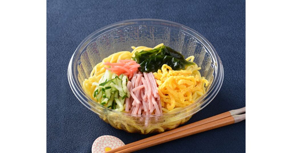 透明な器に盛り付けられた冷やし中華（きゅうり、ハム、錦糸卵、わかめ、紅生姜）と箸のイメージ