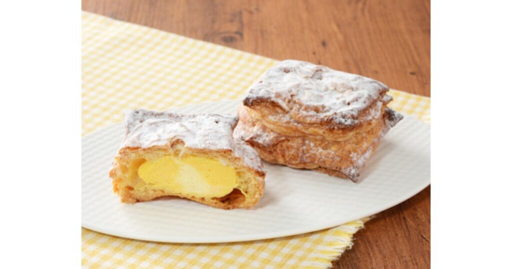 粉砂糖がまぶされた四角いパイ生地のシュークリーム。半分にカットされた断面から、濃厚な黄色のカスタードクリームがたっぷり詰まっている様子がわかる。