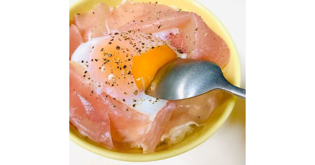 ご飯の上にたっぷりの生ハムと半熟卵を乗せ、黒胡椒を振った丼の写真。スプーンで卵を割り、とろりとした黄身が溢れ出ている様子