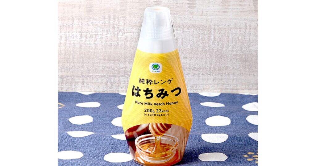 ドット柄の青い布の上に置かれた、ファミリーマートのプライベートブランド（ファミマル）の「純粋レンゲはちみつ」のボトル（200g）。ラベルにはファミマルのロゴ、商品名、カロリー、はちみつが垂れているハニーディッパーのイラストが記載されている。