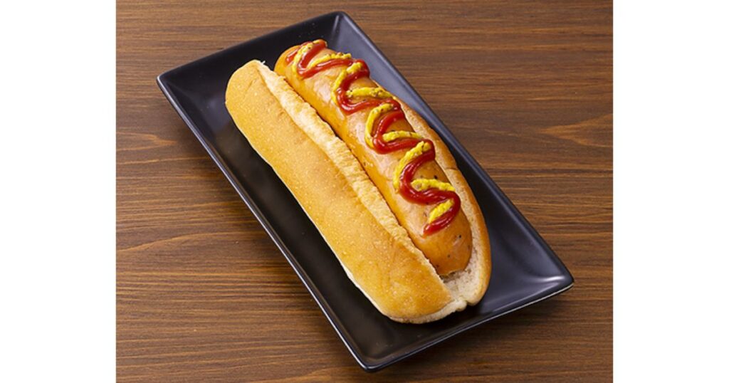 木目調のテーブルの上の黒いお皿に盛り付けられた、粗挽きソーセージのホットドッグ（ローソンの商品イメージ）