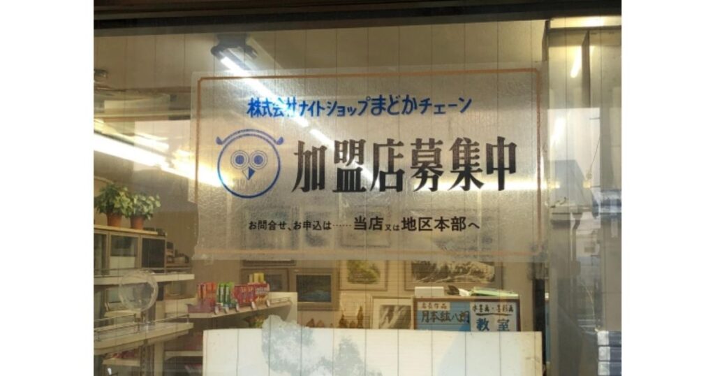 店舗のガラス窓に貼られた「株式会社ナイトショップまどかチェーン 加盟店募集中」と書かれたフクロウのロゴ入り掲示。窓の奥には水墨画が見える