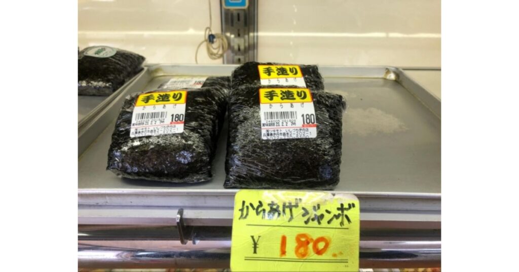 ショーケースのトレイに並べられた、ラップで包まれた大きな手作りの「からあげジャンボ」おにぎり（180円）