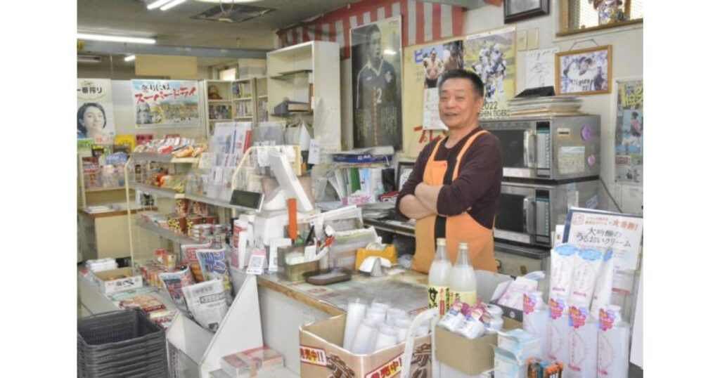 レジカウンター内で腕を組み、笑顔で立つオレンジ色のエプロン姿の男性店主。カウンターには商品や電子レンジが並んでいる