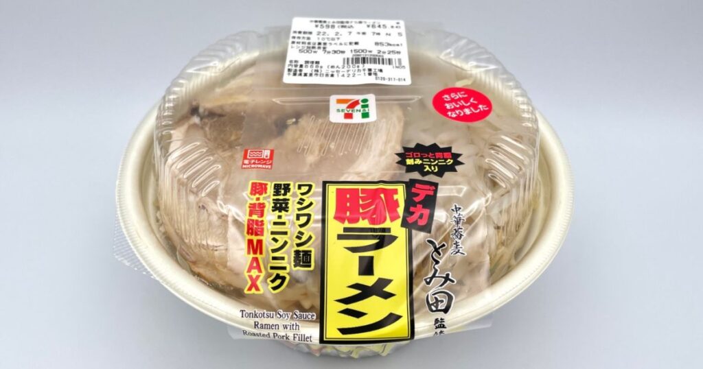 セブン-イレブンのロゴと「中華蕎麦とみ田監修 デカ豚ラーメン ワシワシ麺 野菜・ニンニク 豚・背脂MAX」のステッカーが貼られた、セブン-イレブンのJiro系ラーメンのパッケージ全体写真。価格645.84円（税込）、熱量853kcalの表示あり。