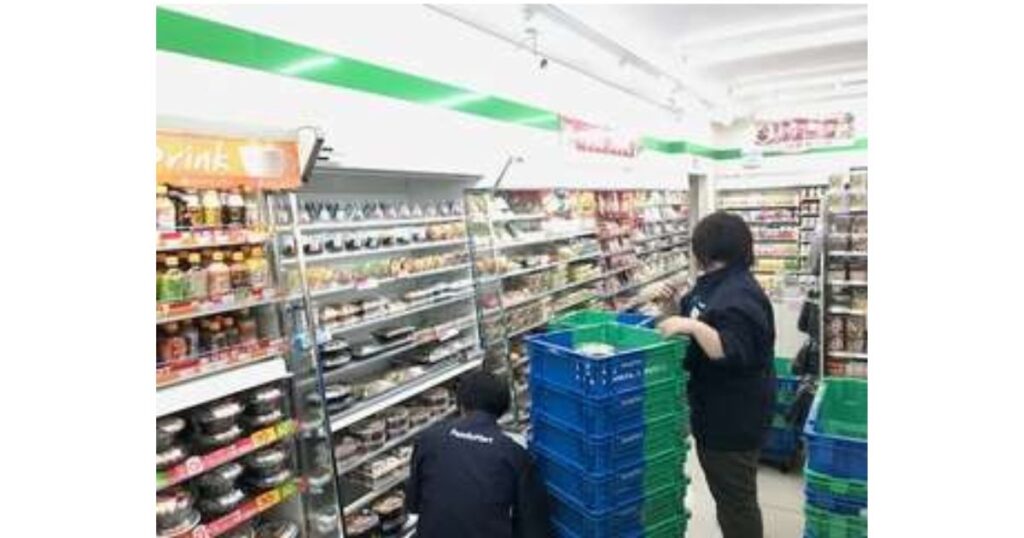 深夜のコンビニ店舗内で、青いコンテナからお弁当や惣菜の品出し作業を黙々と行う2人の店員