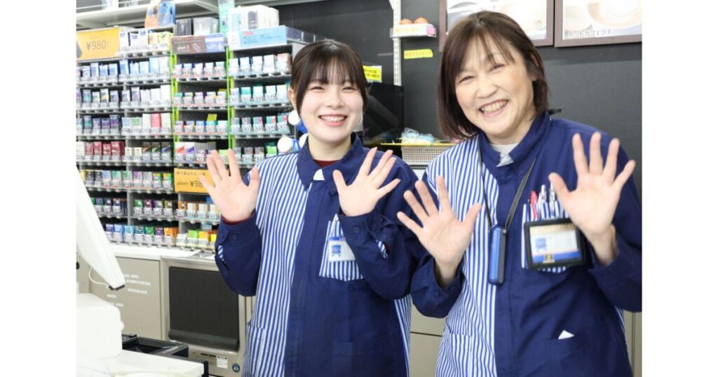 コンビニのレジカウンター内で笑顔で手を振る2人の女性スタッフ