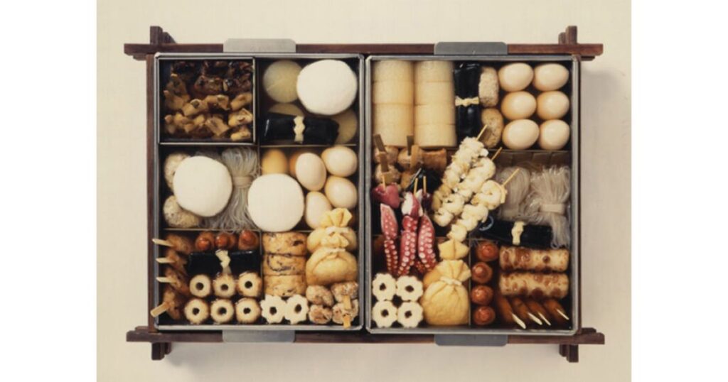 四角い仕切り付きのおでん鍋に、大根、たまご、こんにゃく、ちくわ、たこ串、餅入り巾着など、多種多様なおでんの具材がぎっしりと綺麗に並べられている様子を上から撮影した写真。