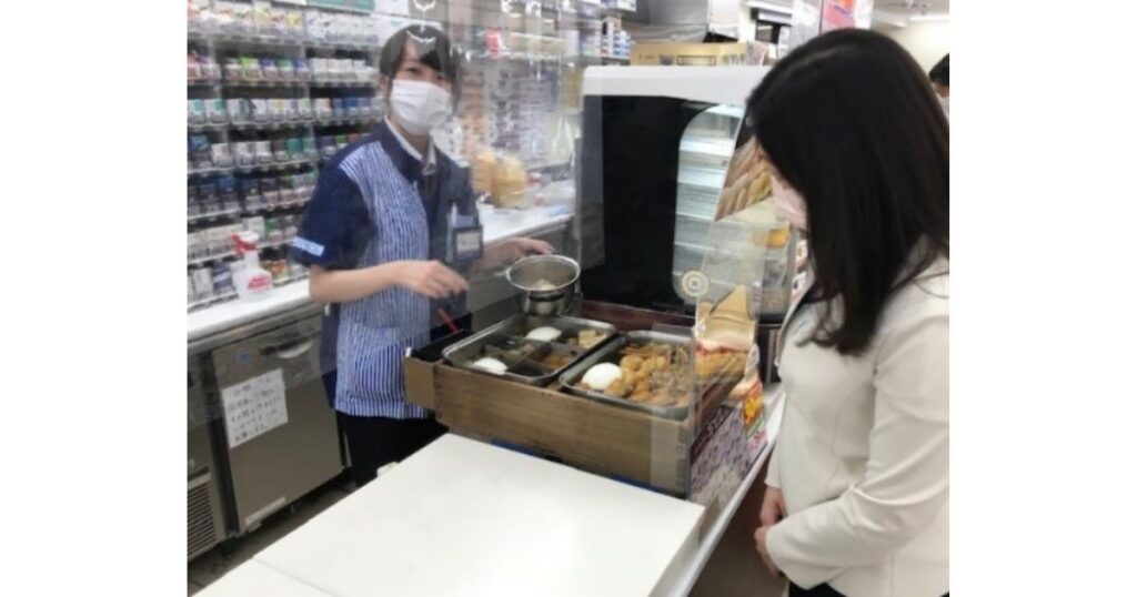 コンビニエンスストアのレジカウンターで、マスクと制服姿の店員が飛沫防止用のアクリル板越しに、女性客へおでんをカップに取り分けて販売している接客風景の写真。