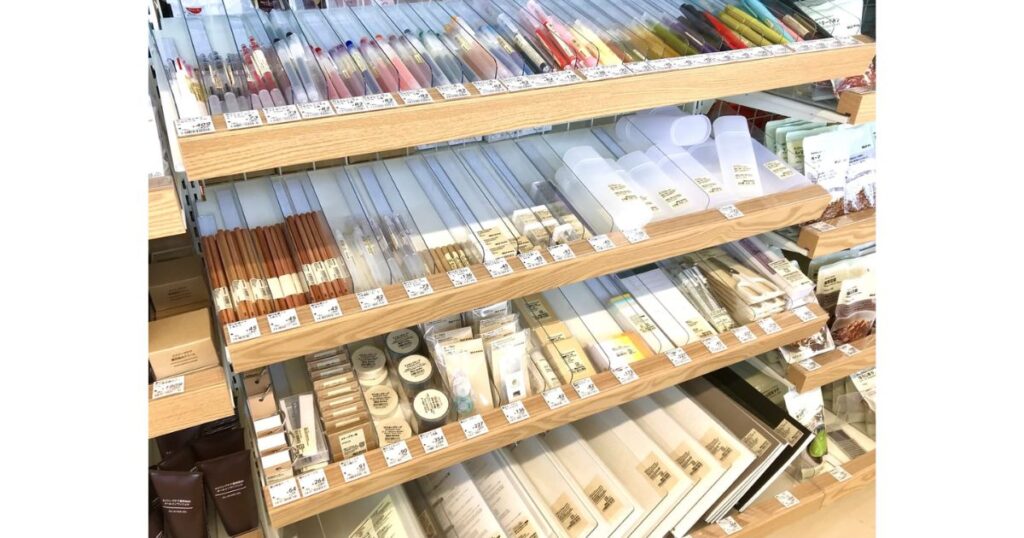 コンビニエンスストアまたは文房具店の陳列棚。木製の棚板に無印良品の木目調鉛筆や様々なペン、ノートなどの文房具が整理されて並んでおり、それぞれの商品にプライスタグが付いている。棚の奥にはレジカウンターと店員が見える。