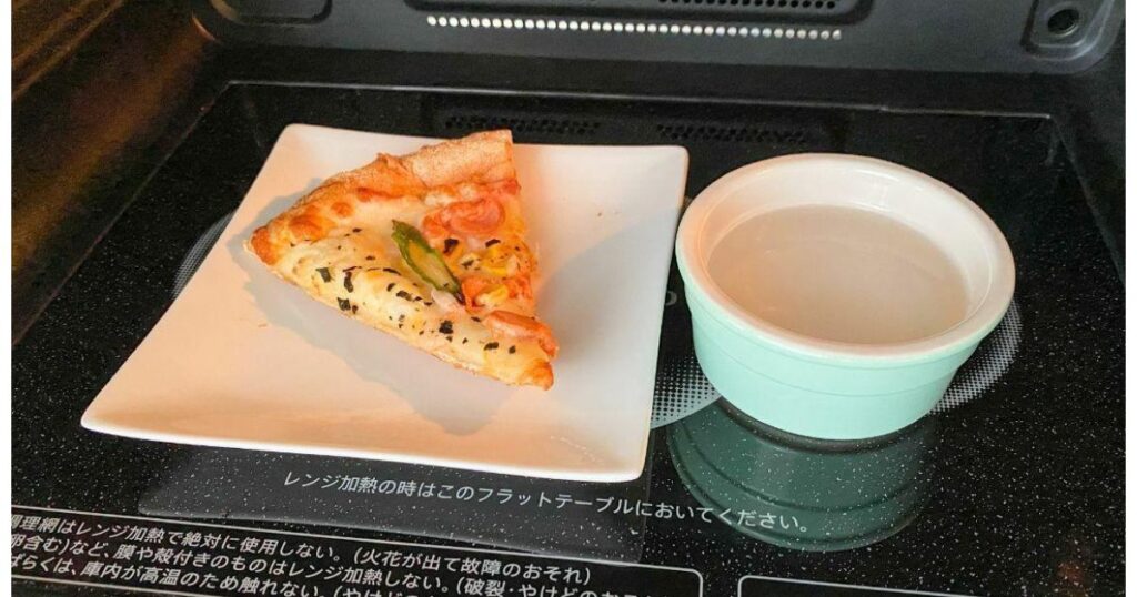 電子レンジ庫内に置かれた1切れのピザと、美味しく温めるために水を入れた小さな耐熱容器