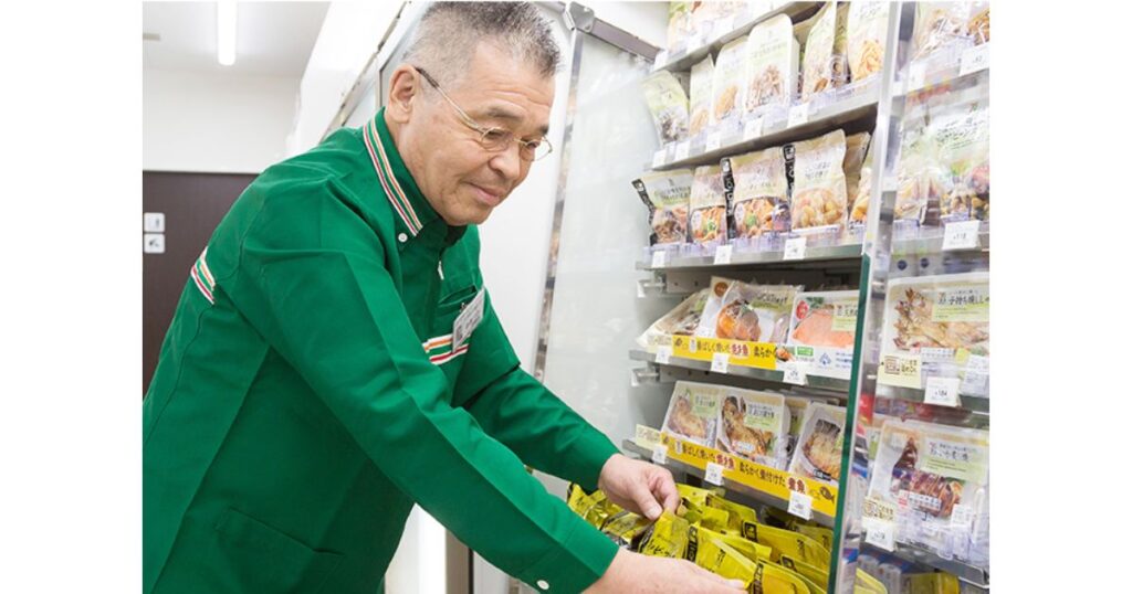 セブン-イレブンの制服を着て、チルド惣菜の棚を整理するシニア男性店員の写真