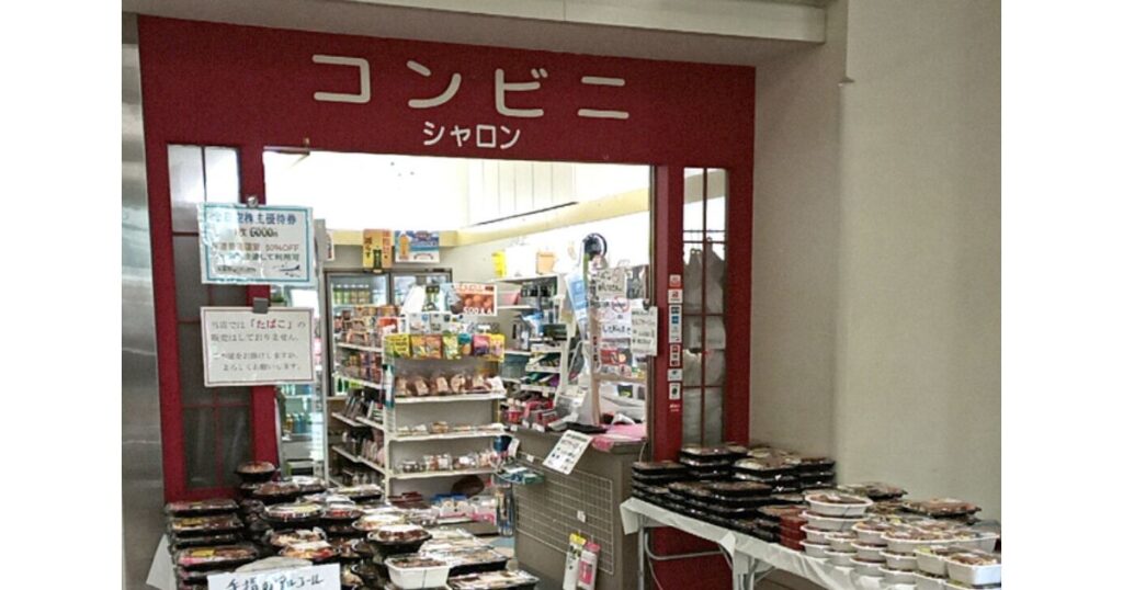 「コンビニ シャロン」の看板が掲げられた実在する店舗の入り口と、店先に山積みされた手作りのお弁当