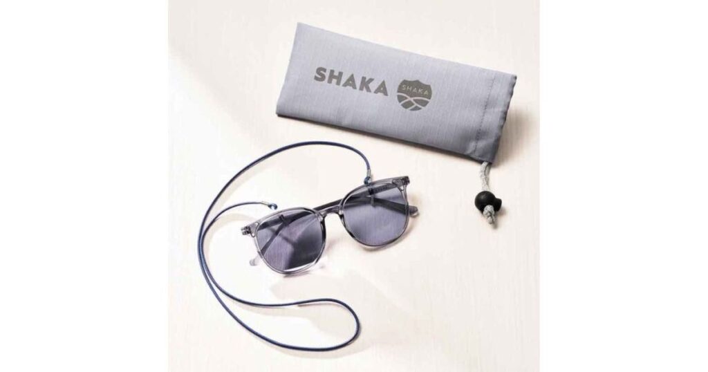 白い背景に置かれたクリアグレーのサングラス。青いグラスコード(紐)が装着されており、上部には「SHAKA」のロゴが入ったグレーの専用ポーチが添えられている。