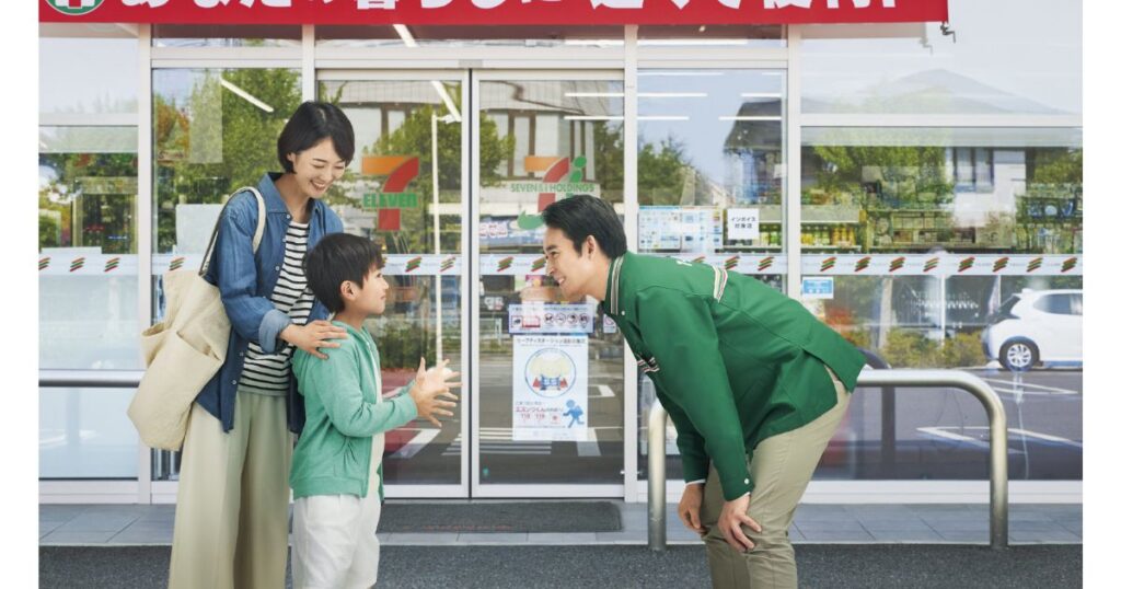 「セブン-イレブンの店舗前で、緑色の制服を着た店員が親子連れに笑顔で挨拶をしている写真。」