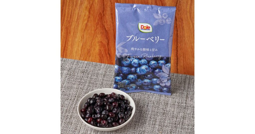 木目調のテーブルの上に置かれたグレーのランチョンマットの上に、Dole（ドール）の冷凍ブルーベリーのパッケージが立っている。その手前には、白いボウルに山盛りの冷凍ブルーベリーが入っている。