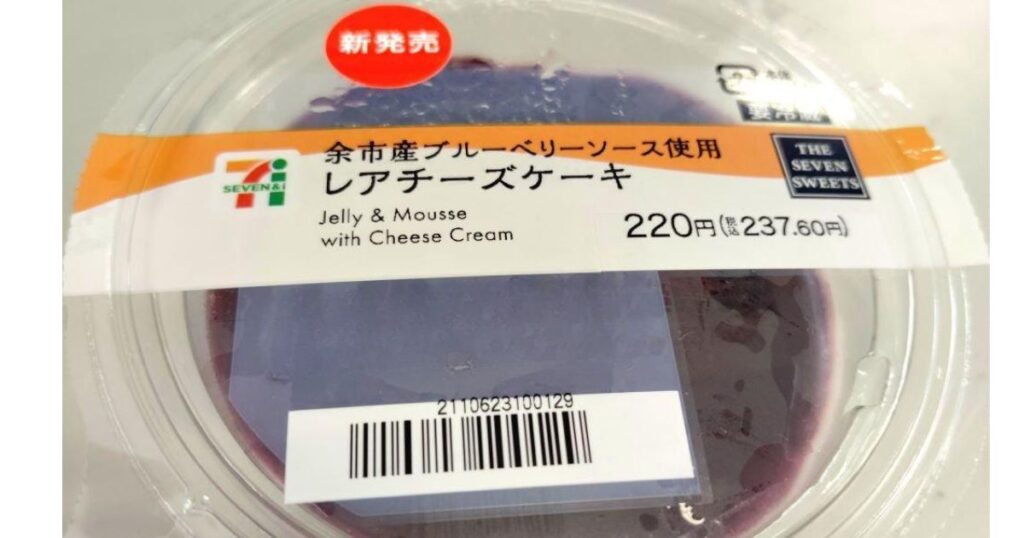 セブンプレミアムの「余市産ブルーベリーソース使用 レアチーズケーキ」のプラスチック容器。蓋には商品名、価格、バーコードが印刷されたラベルが貼られており、赤い「新発売」のシールが付いている。蓋越しに紫色のブルーベリーソースが見える。