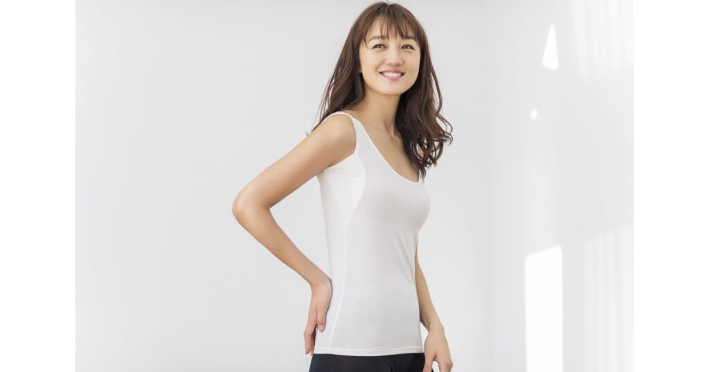白い背景の前で、白いキャミソールを着用して腰に手を当てて立つ女性。
