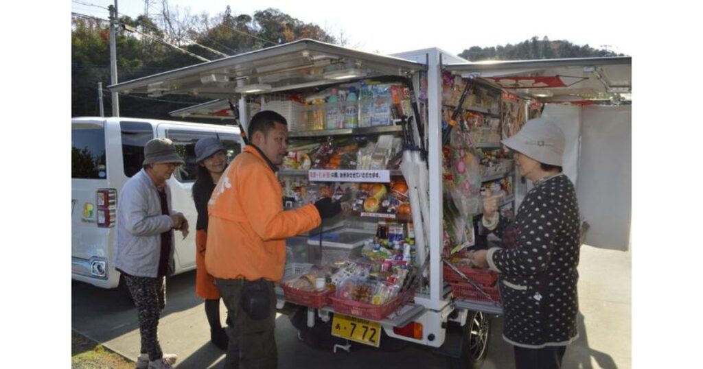 住宅街で移動スーパー(とくし丸など)を利用して買い物をする地域住民とスタッフの様子。コンビニ閉店後の買い物難民対策や、対面販売による高齢者の見守りといったラストワンマイルの解決策を示している。