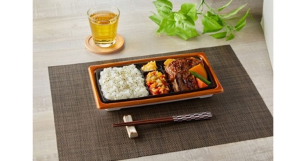 ランチョンマットの上に綺麗に並べられた、ハンバーグや副菜、白飯が入ったコンビニ弁当と冷たいお茶のセット。