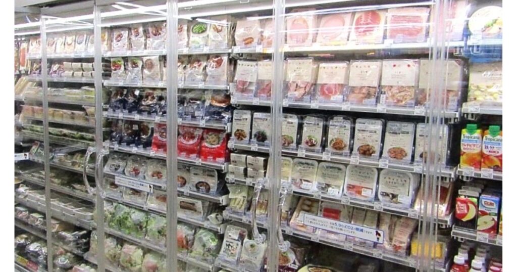コンビニのチルド食品コーナー。ガラス扉の棚に豆腐、惣菜、ドリンクなどが並び、TOPVALUの餃子の皮も見られる。