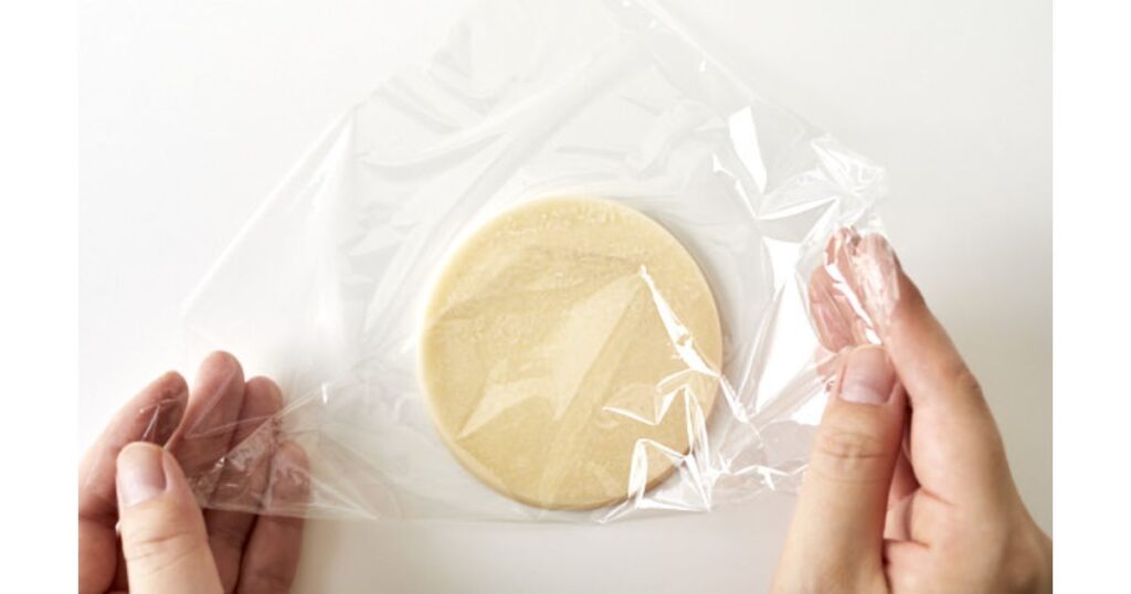 ラップで包まれた丸い餃子の皮の束。人の両手がラップの端を整えている。