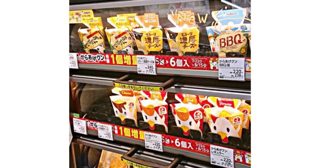 ローソンのレジ横にあるホットスナック用ショーケース内の写真。「からあげクン」の様々なフレーバーのパッケージが棚に並んでいます。上段には期間限定の「濃厚チーズ味」や「BBQ味」、下段には定番の「からあげクン レッド」や「レギュラー」が配置されています。それぞれの棚には「1個増量(6個入)」という赤いセールスプロモーションのポップが貼られています。