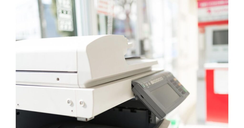 明るい店舗内に設置された大型マルチコピー機の斜め前からのクローズアップ写真。上部の原稿台と操作パネルが写っており、背景は白くぼかされています。