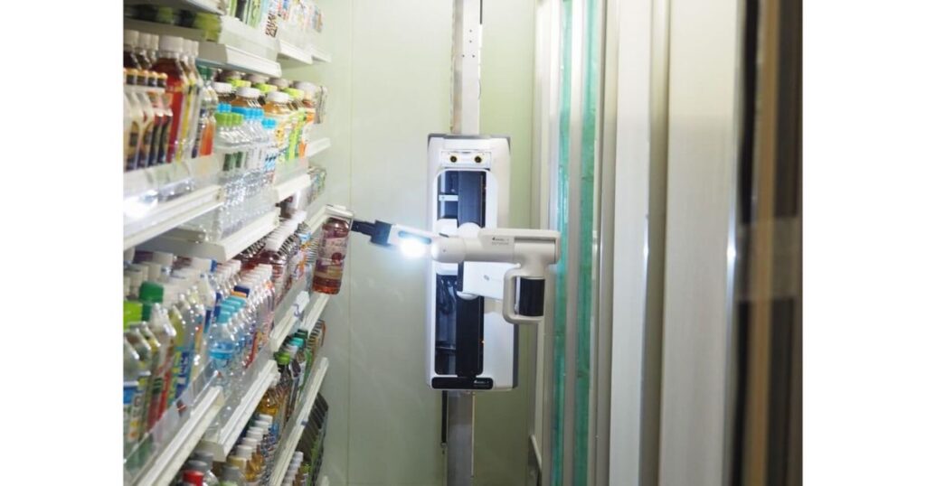 コンビニエンスストアのクールケース内で動作する自律型ロボットのクローズアップ写真。白いボディに青いアクセントがある。ロボットのアームは先端が光っており、ボトルを掴んでいる。アームは棚に積まれた様々なペットボトル（主に飲料）の列に向けて伸びている。