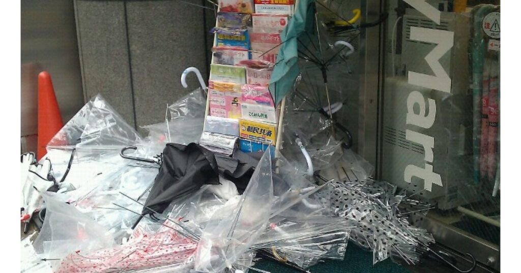 コンビニエンスストアの入り口付近に、無造作に山積みにされた大量の放置傘の写真。