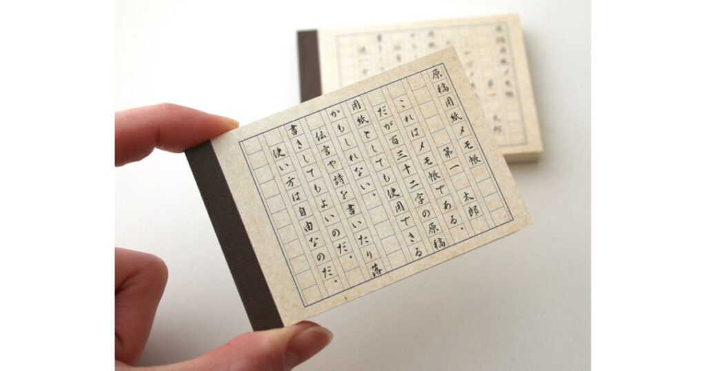 手のひらサイズの小さな原稿用紙風メモ帳に、実際に縦書きで文章が書き込まれている様子。