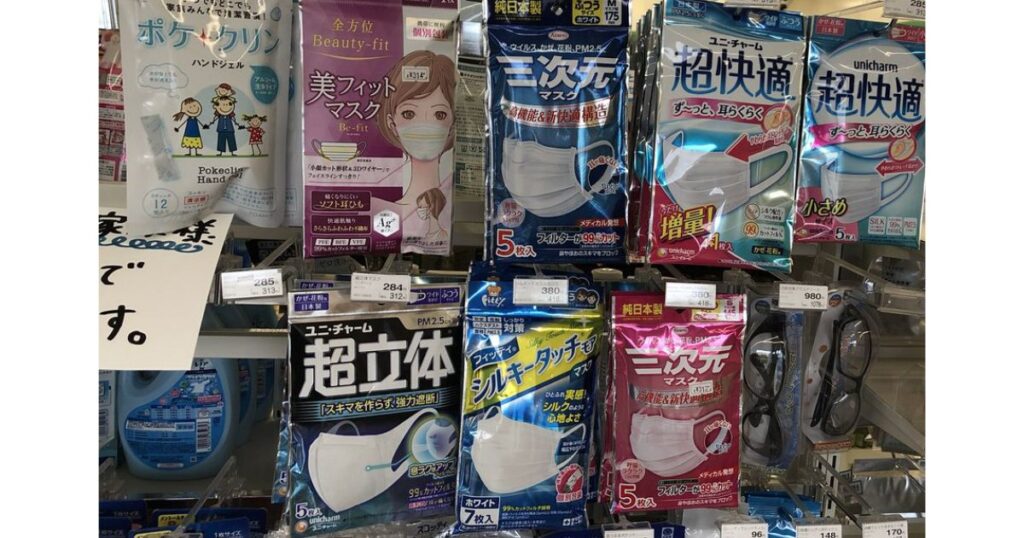 実際の写真。コンビニのマスク売り場の棚に、「美フィットマスク」「三次元マスク」「超快適」「超立体」など様々な種類のマスクが陳列されており、それぞれに価格タグが付いている。