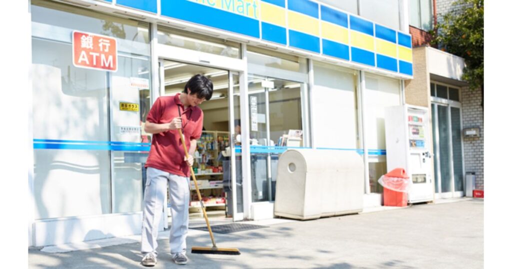 晴れた日に FamilyMart 店舗の前で、赤い制服を着た男性店員がホウキで歩道を掃いている実写画像。ガラス窓には「銀行 ATM」のステッカーが見える。