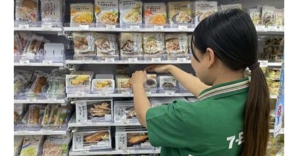 コンビニのチルド惣菜コーナーで、緑色の制服を着た女性店員が商品を棚に陳列している後ろ姿。棚には多くのサラダやお惣菜が並んでいる。