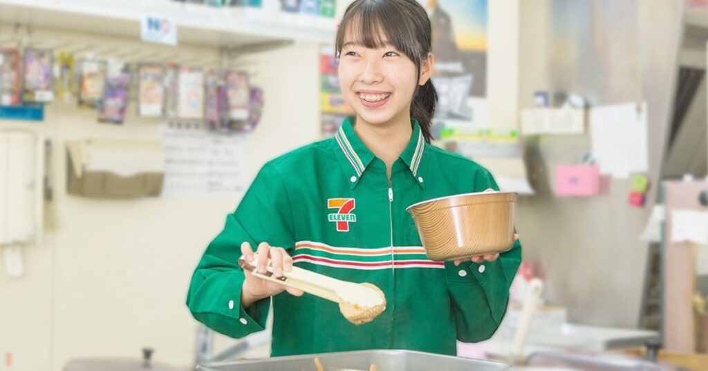 7-ELEVEn の制服を着た笑顔の女性店員が、レジカウンターでトングを使ってフライドチキンをパックに入れている実写画像。