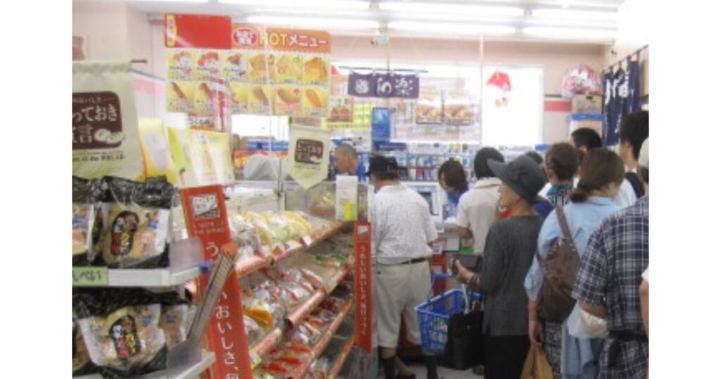 コンビニ店内で、レジカウンターの前に長い列を作る多くの客の後ろ姿。店内は混雑しており、上部には「HOT メニュー」の大きなポップが見える。