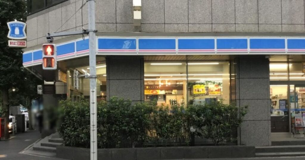 街の角地にあるローソン（LAWSON）店舗の外観を捉えた写真。グレーの石造りの建物で、青と白のストライプのパラペット看板が角に沿って伸びている。左端には、青地に白いミルク缶が描かれた丸いLAWSONのロゴ看板と、その下に「たばこ 銀行ATM」と書かれた看板が設置されている。その隣には赤信号を点灯させた歩行者用信号機がある。大きなガラス窓からは明るい店内が見える。歩道には緑の植栽が並んでいる。