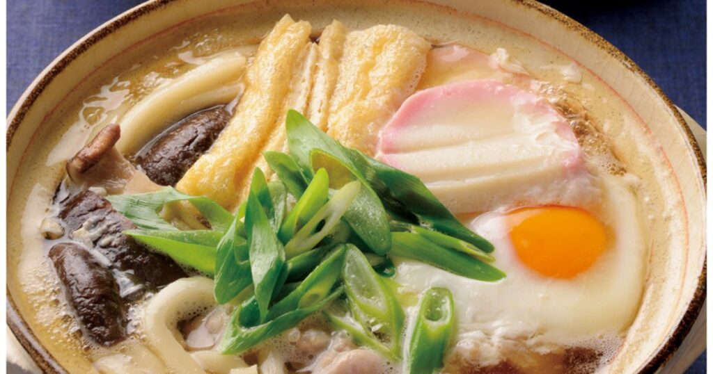 土鍋でぐつぐつと煮込まれる、卵、ネギ、油揚げ、椎茸、かまぼこなどの具材がたっぷり乗った鍋焼きうどんのアップ