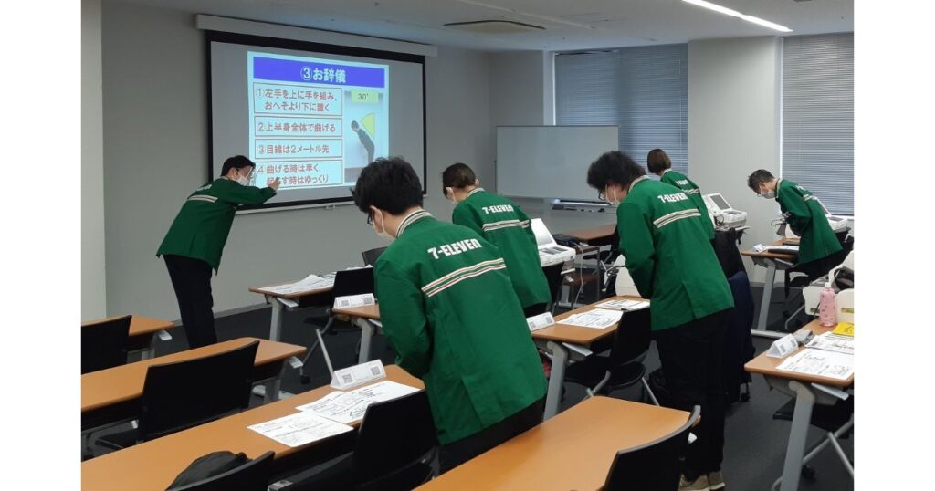 セブン-イレブンの制服を着た従業員たちが、研修室でプロジェクターに映し出された指示を見ながら、お辞儀の練習をしている様子。