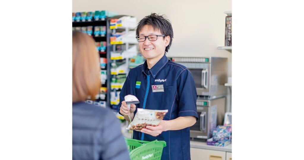 ファミリーマートの制服を着た男性店員が、笑顔でレジ接客をしている様子。