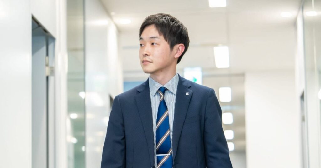 明るいオフィスの廊下を歩く、紺色のスーツを着た若い男性ビジネスマン（本部社員や店長のイメージ）の写真