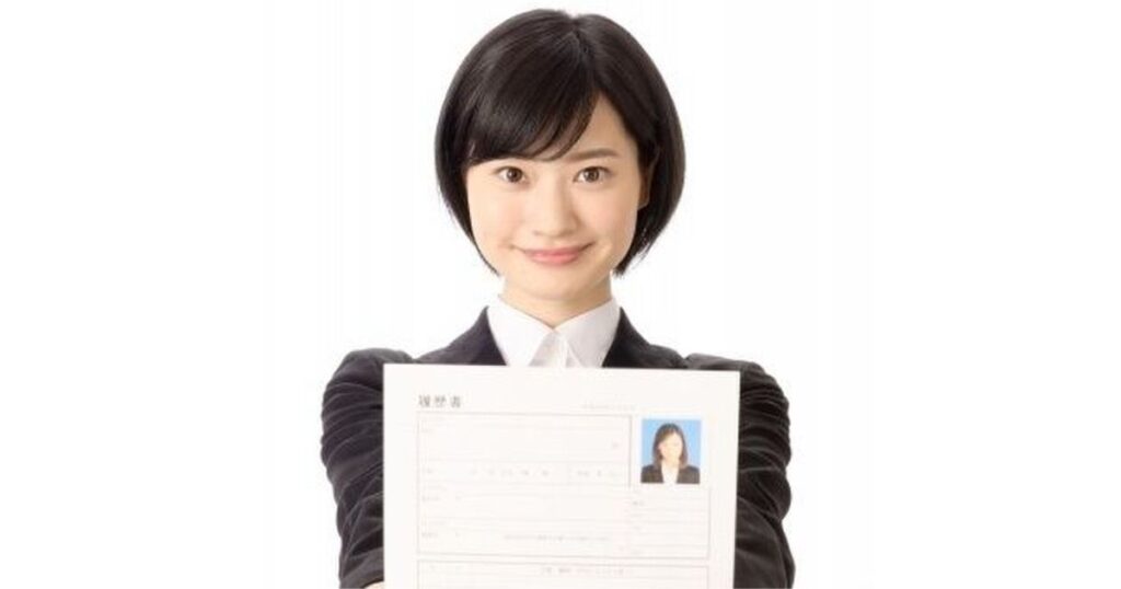 リクルートスーツを着た女性が、証明写真を貼った履歴書を両手で持ち、カメラに向かって提示している様子。