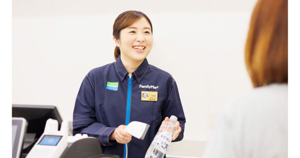 ファミリーマートの青い制服を着用した女性店員が、レジカウンターで商品のバーコードをスキャンしながら笑顔で対応している場面。