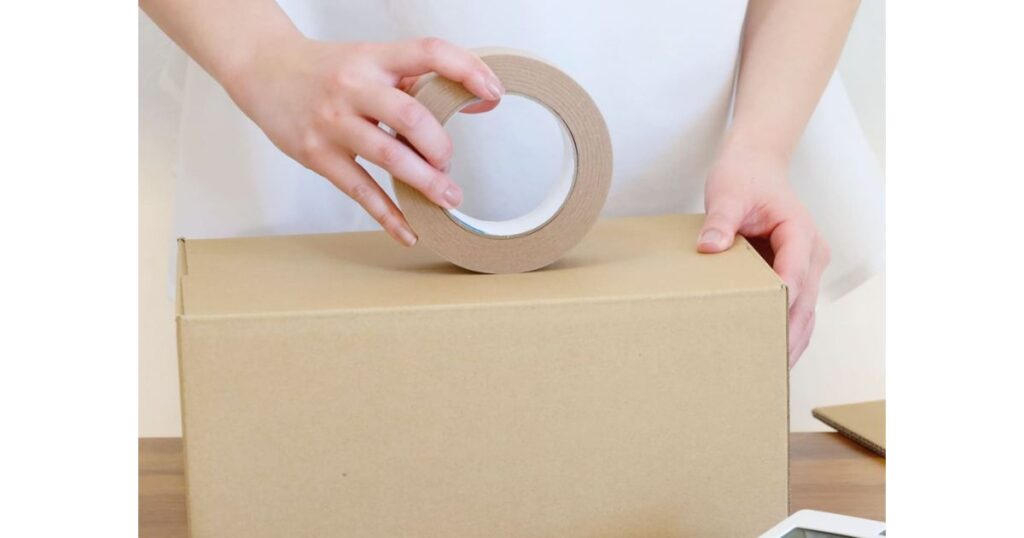 組み立てられた茶色の無地ダンボール箱の上面に、両手でクラフトテープを貼って梱包している様子。