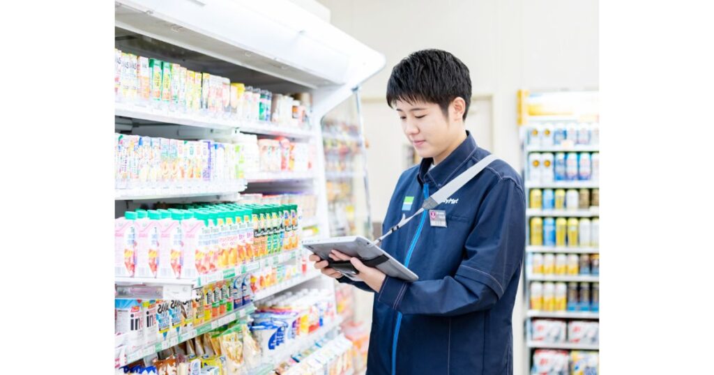 コンビニの店内で、タブレット端末を手に持ちながら真剣な表情でチルド棚の飲料商品を検品・陳列している制服姿の男性店員の写真。