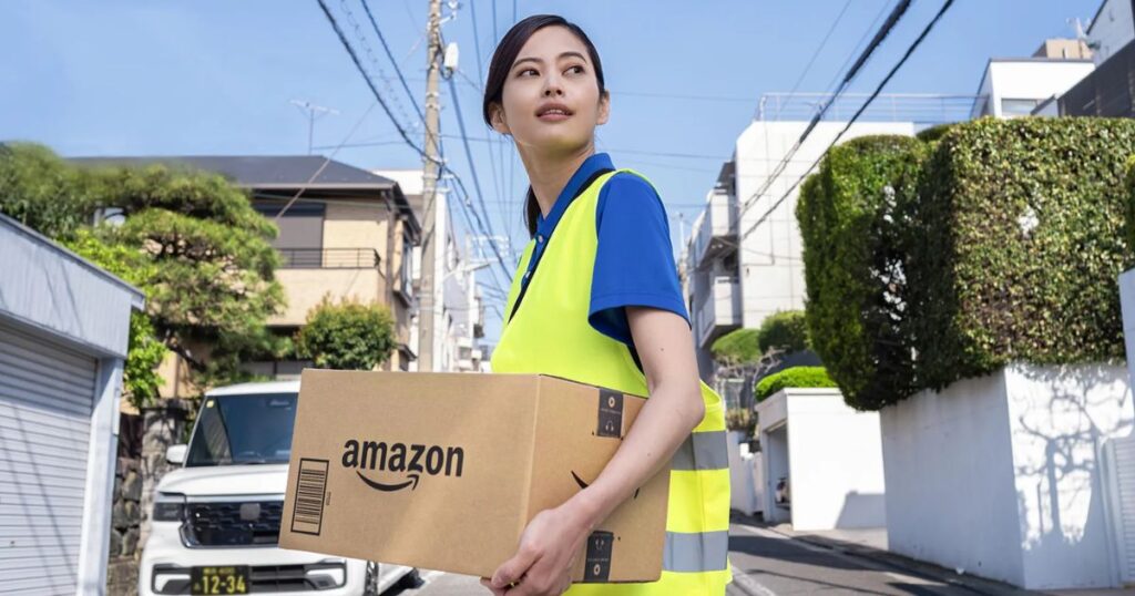 住宅街の道路で、Amazonのロゴが入った段ボール箱を抱えて歩く、黄色いベストを着た女性配達員の写真。